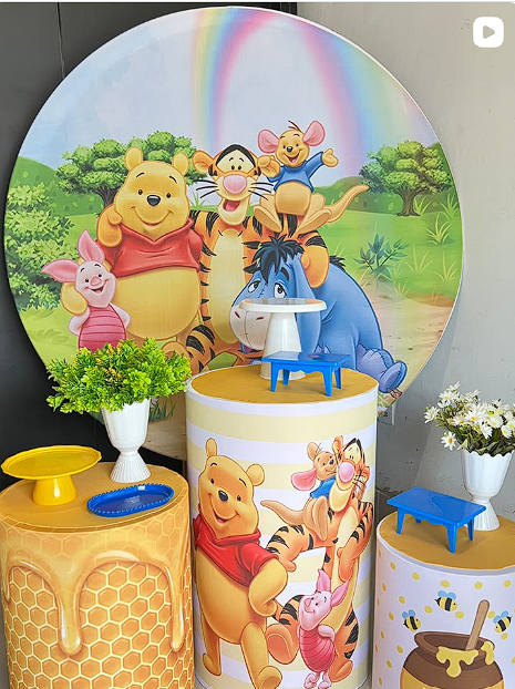 Cenário de festa infantil com painel circular, cilindros decorativos, flores e suportes para doces