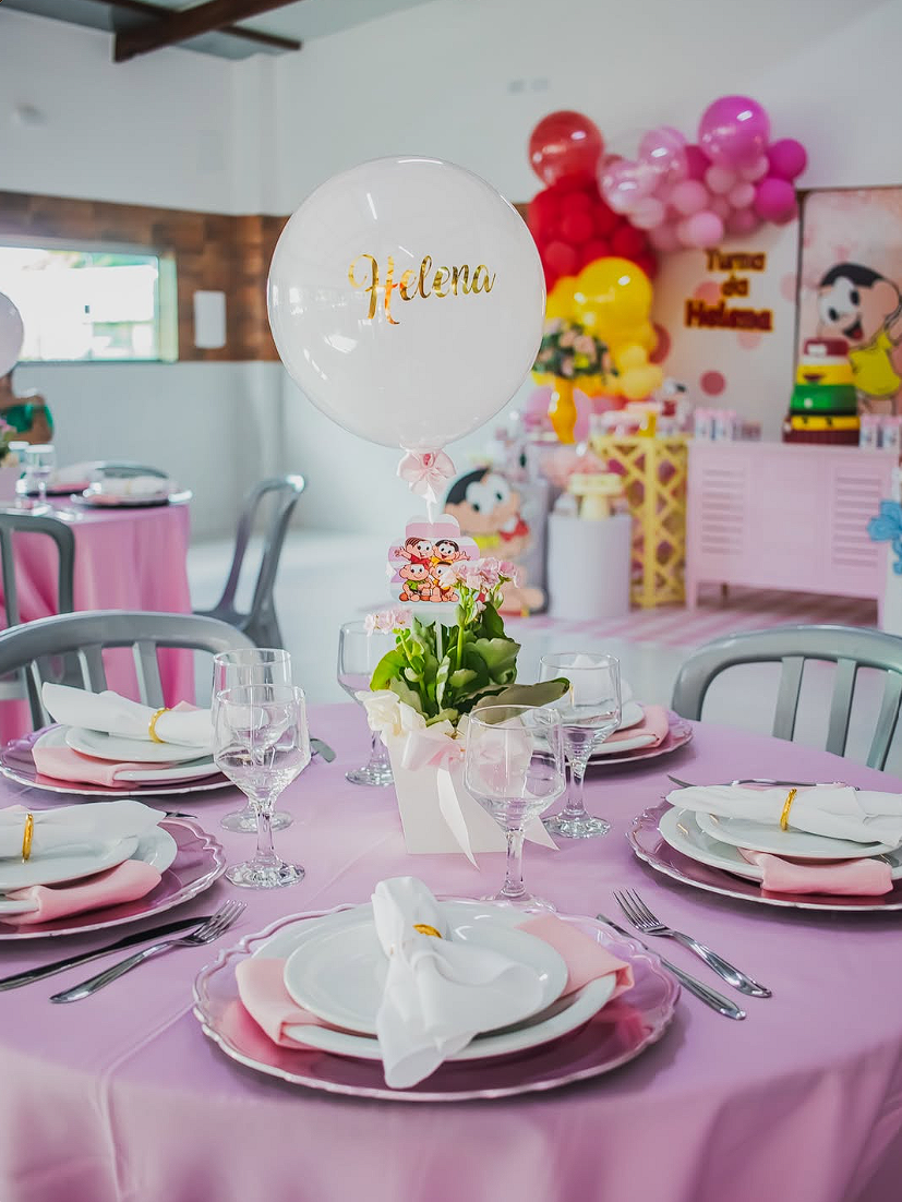 Mesa de aniversário infantil em tons de rosa com louças, taças, flores e balões ao fundo
